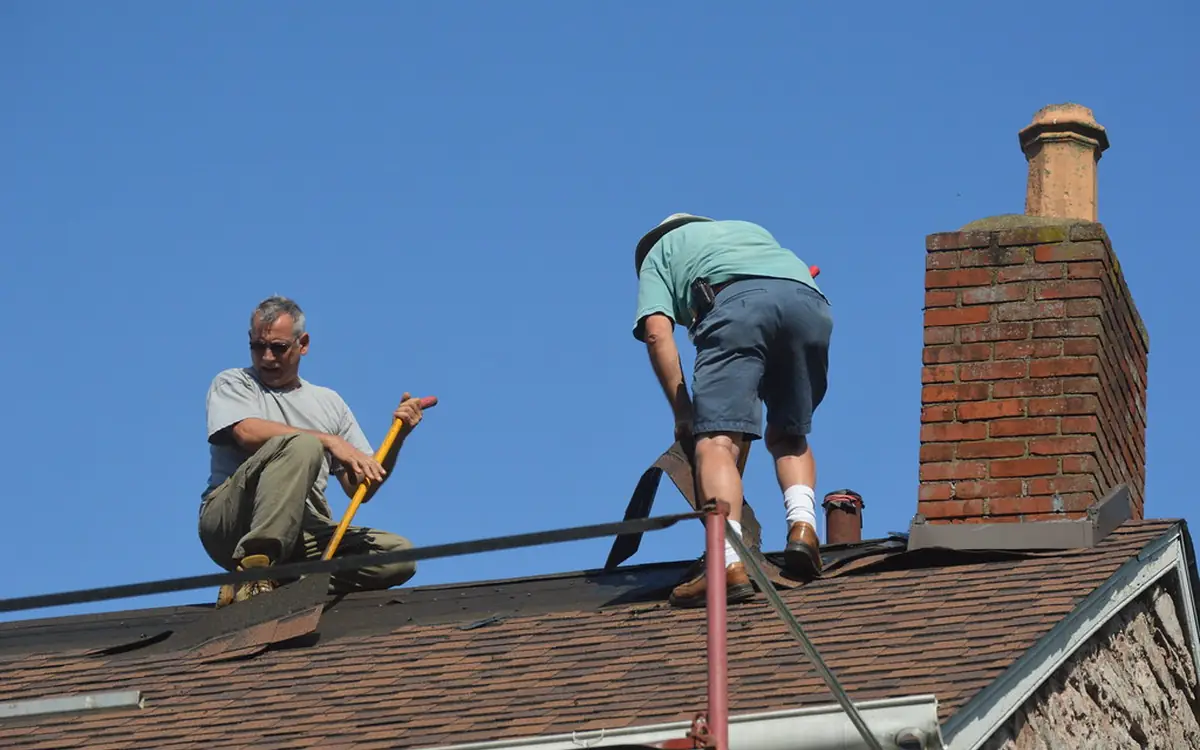 Two roofers working on an asphalt shingle roof next to a brick chimney