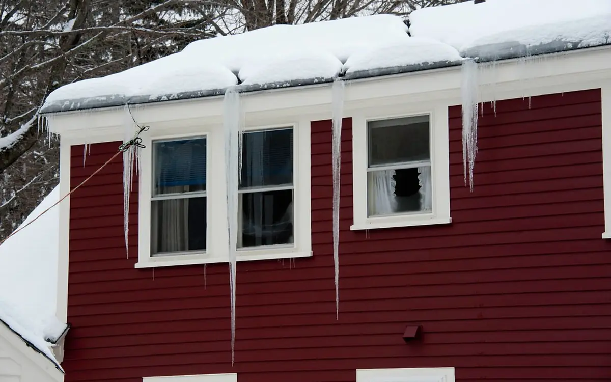 Ice dam and long icicles hanging from the snowy roof edge of a red house