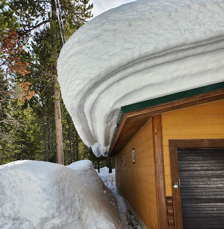 Thick snow cornice curling several feet off a wooden building's roof in a subarctic climate, with visible horizontal layering typical of heavy mountain snowpack