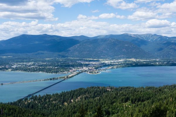 Wide view across Lake Pend Oreille toward Sandpoint, with a long railroad trestle and adjacent vehicle bridge spanning the water, backed by forested Cabinet and Selkirk mountains