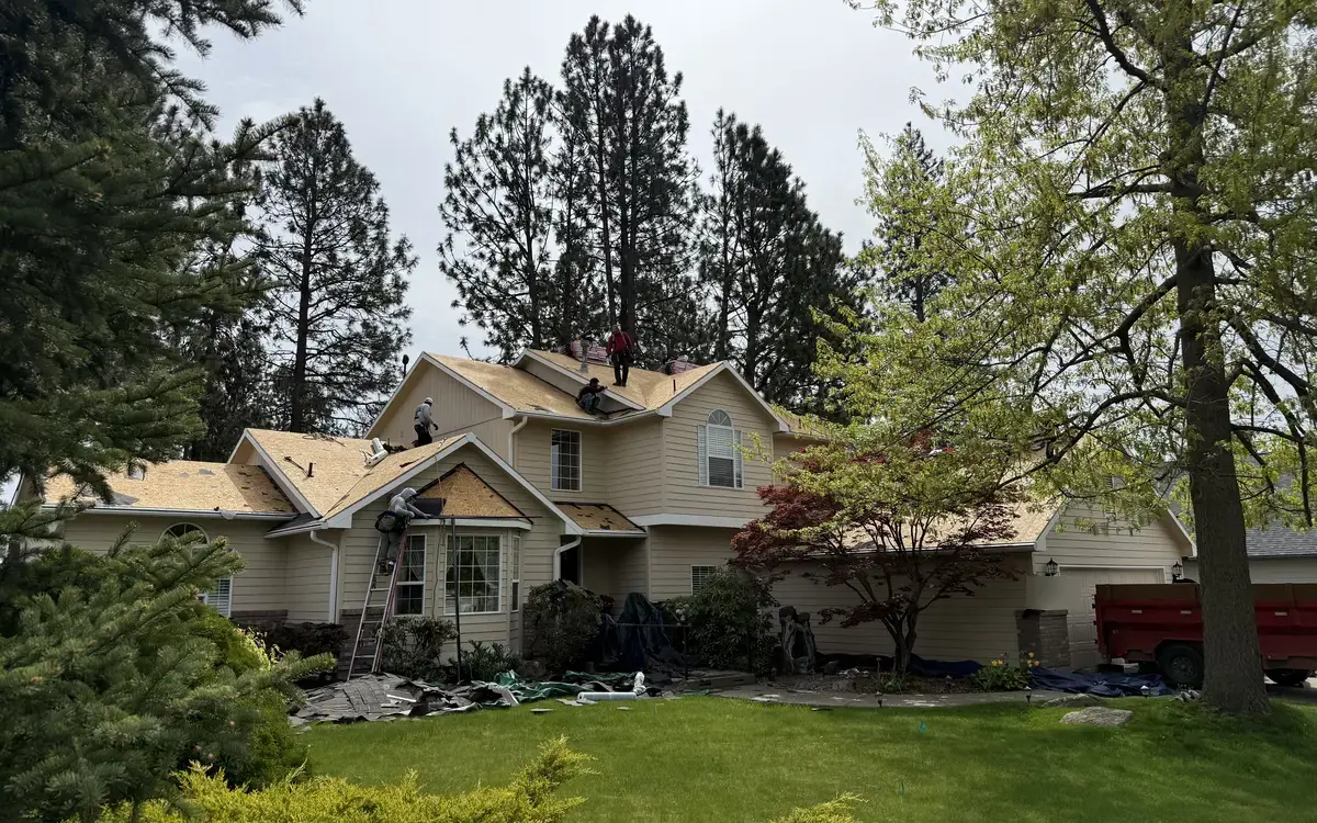 Roofing crew tearing off old shingles down to the roof deck on a two-story house, with tarps over the landscaping and a dump trailer in the driveway