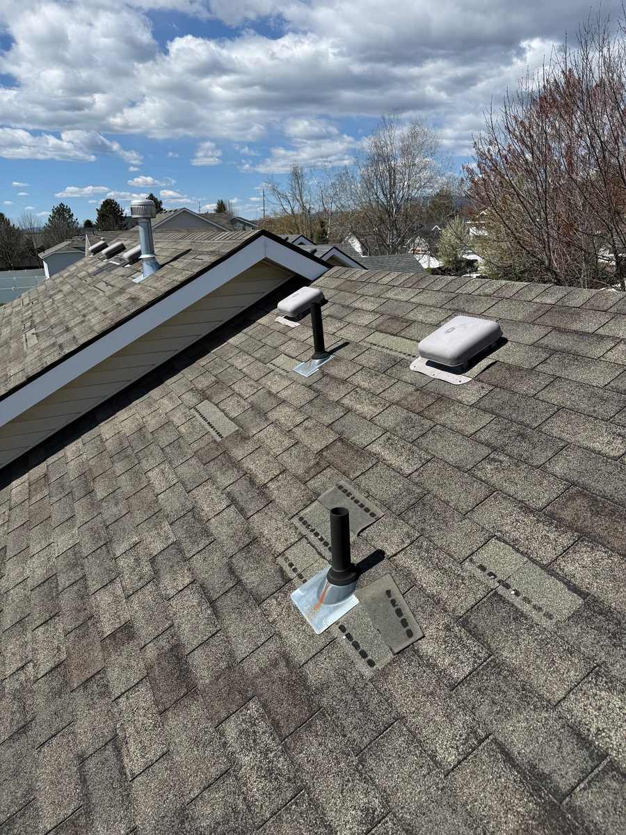 Asphalt shingle roof showing multiple creased and lifted shingle tabs near a plumbing vent pipe from wind damage, with visible granule loss